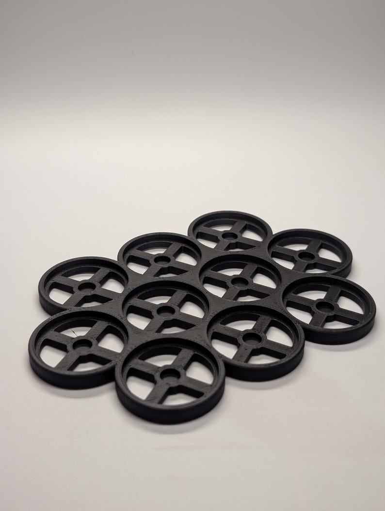 May include: A cluster of eleven black, circular coasters with a wheel-like design. Each coaster has a central hub with spokes extending to the outer rim. The coasters are arranged in a honeycomb pattern, suitable for holding multiple glasses or mugs.
