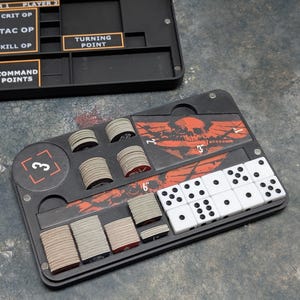 Może przedstawiać: Czarny organizer do gier z kośćmi, żetonami i kartami. Organizer posiada przegródki na elementy gry i zawiera tekst, np. "Turning Point" oraz wymiary w calach (6" to ok. 15 cm).