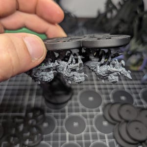 May include: A close-up of a miniature model with a circular base, featuring detailed figures and a textured surface. The model is held by a hand, with additional circular bases and other miniature components visible in the background.