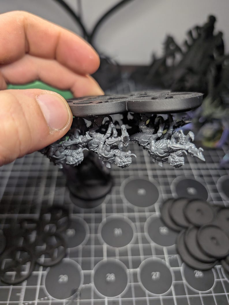 May include: A close-up of a miniature model with a circular base, featuring detailed figures and a textured surface. The model is held by a hand, with additional circular bases and other miniature components visible in the background.