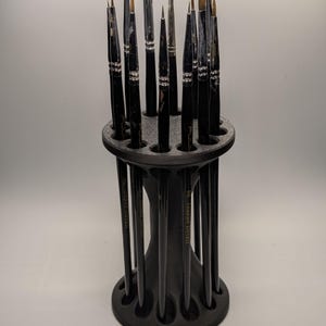 May include: A black paint brush holder filled with various paint brushes. The holder has a circular top and base with a curved, hourglass-shaped body. The paint brushes are of different sizes and shapes, with black handles and silver accents.