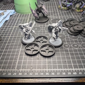 May include: Miniature figurines of winged warriors with swords and guns, along with black circular bases and other components, are arranged on a black cutting mat. Additional figurines and a green container are visible in the background.