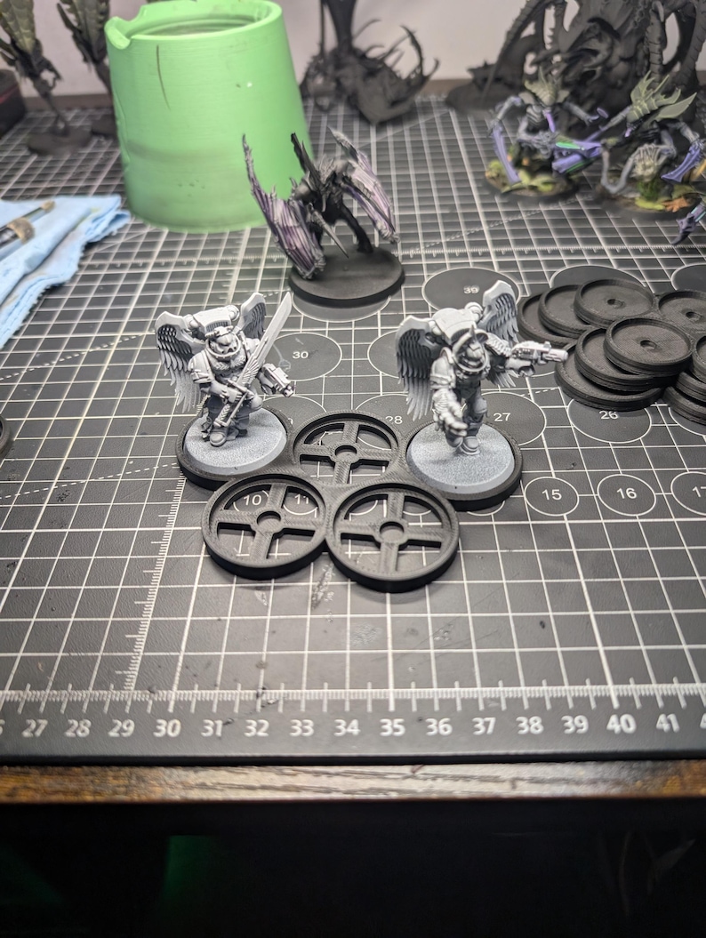 May include: Miniature figurines of winged warriors with swords and guns, along with black circular bases and other components, are arranged on a black cutting mat. Additional figurines and a green container are visible in the background.