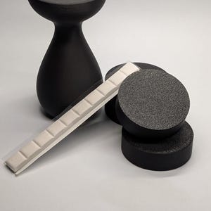 May include: A black, matte-finish candle holder with a circular top and a rounded base. Beside it are three black, textured, circular objects and a strip of small, white, square tiles.