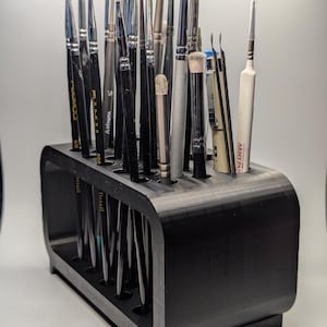 Peut inclure: Un porte-pinceaux noir imprimé en 3D rempli de divers pinceaux. Le support comporte plusieurs fentes pour organiser les pinceaux de différentes tailles et formes. Les pinceaux ont des manches noirs et des couleurs de poils variées, dont le blanc, le marron et le gris.