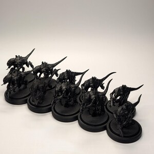 May include: A collection of black miniature figurines, possibly for tabletop gaming. The figures are detailed, with insectoid or reptilian features, and are mounted on round black bases. The lighting is bright, highlighting the textures.