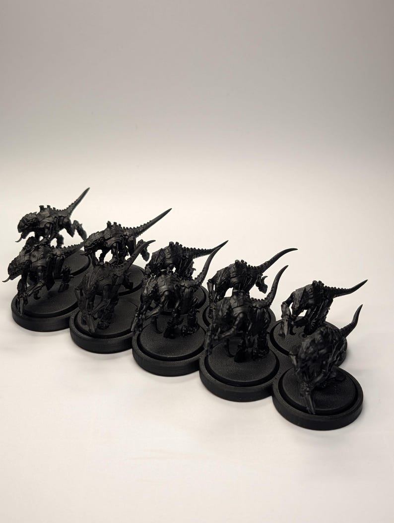 May include: A collection of black miniature figurines, possibly for tabletop gaming. The figures are detailed, with insectoid or reptilian features, and are mounted on round black bases. The lighting is bright, highlighting the textures.