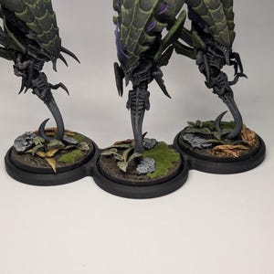 May include: Three detailed miniature figures of a creature with a segmented, spiky carapace in shades of green and purple. The figures stand on a black base with miniature terrain, including rocks and foliage. The figures have a skeletal appearance.