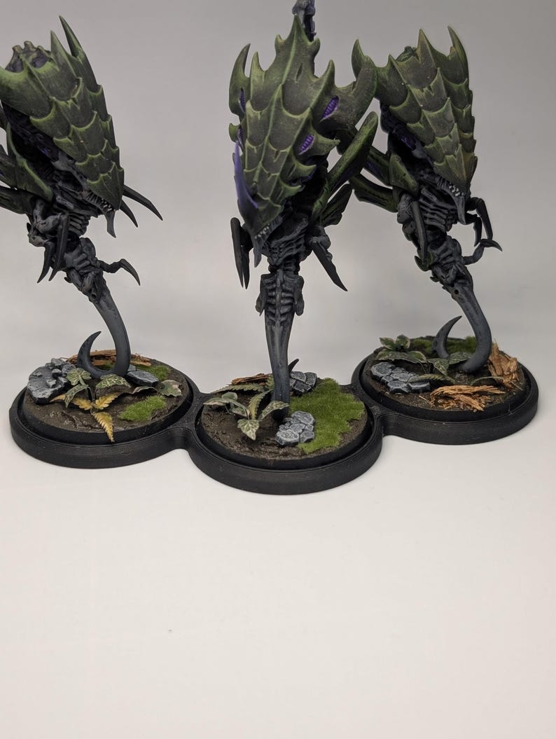 May include: Three detailed miniature figures of a creature with a segmented, spiky carapace in shades of green and purple. The figures stand on a black base with miniature terrain, including rocks and foliage. The figures have a skeletal appearance.