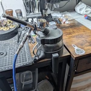 Puede incluir: Un sistema de aerógrafo con un recipiente negro y un aerógrafo plateado. Una manguera de aire azul está conectada al aerógrafo. El aerógrafo está sobre una mesa de madera con otros suministros de arte.