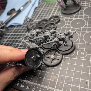 May include: Miniature figurines of insect-like creatures, unpainted, on black bases. The figures are held in place by a magnetic base. A partially painted winged creature stands in the background. A paintbrush and a blue cloth are also visible.
