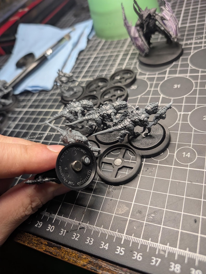 May include: Miniature figurines of insect-like creatures, unpainted, on black bases. The figures are held in place by a magnetic base. A partially painted winged creature stands in the background. A paintbrush and a blue cloth are also visible.