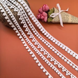 Puede incluir: Tres cintas de encaje de algodón blanco con una fila de pequeños pompones a lo largo del borde. El encaje es delicado y tiene un borde festoneado.