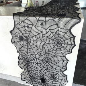 Pode incluir: Caminho de mesa de renda preta com um design de teia de aranha. O caminho de mesa apresenta uma borda ondulada e várias aranhas decorativas. Este caminho de mesa com tema de Halloween é perfeito para adicionar um toque assustador.