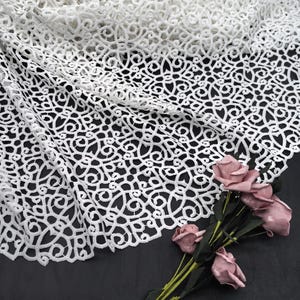 Puede incluir: Tela de encaje blanco con un intrincado patrón arremolinado. El delicado material está drapeado, revelando el detallado diseño calado. Un ramo de rosas rosas con tallos verdes descansa sobre una superficie oscura, complementando el encaje.