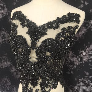 May include: Black beaded lace applique with intricate floral and scrollwork designs. The applique features a sheer mesh backing and is heavily embellished with black beads and sequins, creating a textured, ornate look. Ideal for embellishing garments.
