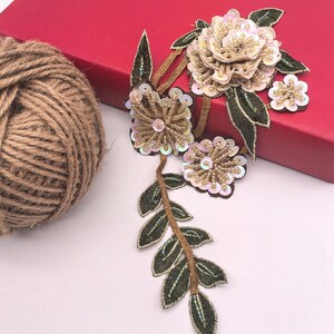 May include: Embroidered floral applique with gold, beige, and green accents. The design features multiple flower shapes with sequin and bead embellishments, along with green leaves and stems. The applique is likely for decorative purposes.