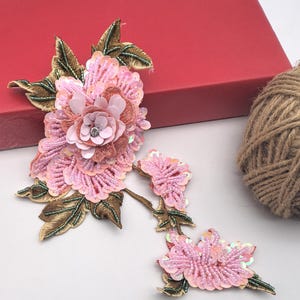 May include: Pink sequined floral appliques with embroidered leaves. The appliques feature a layered design with a central rhinestone. The appliques are set against a red box and a ball of twine.