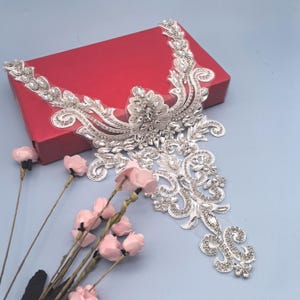 Puede incluir: Aplicación de pedrería plateada y transparente con un diseño floral ornamentado. La aplicación está sobre un respaldo de malla transparente y presenta una variedad de pedrería de diferentes tamaños. Sobre una caja roja con flores rosas en primer plano.