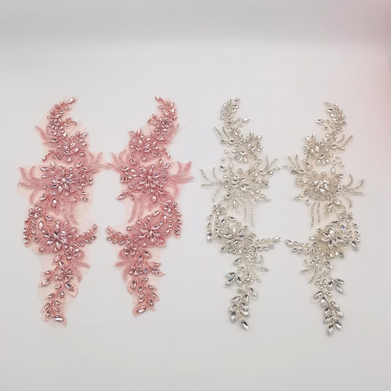 K&ouml;nnte beinhalten: Zwei Paare von Perlen-Applikationen, eine in Rosa und eine in Silber. Jede Applikation zeigt ein florales Design mit kristallartigen Perlen.