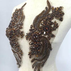 May include: Two ornate, beaded appliques in a floral design. The appliques feature brown, gold, and silver beads and sequins, arranged on a sheer, beige backing. The design is symmetrical, with each applique resembling a stylized wing or feather.