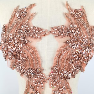 Könnte beinhalten: Ein Paar symmetrische Applikationen in Roségold mit facettierten Kristallen und Perlen in Blumen- und Blattmustern auf einem transparenten, pfirsichfarbenen Netzstoff. Das Design umfasst aufwendige Perlenstickereien und metallische Fäden.