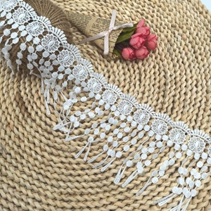 Peut inclure: Une bordure en dentelle blanche avec un motif floral et à pois. La bordure présente un bord festonné et des franges pendantes. Un rouleau de ficelle et un petit bouquet de fleurs roses sont en arrière-plan, posés sur un tapis tissé.