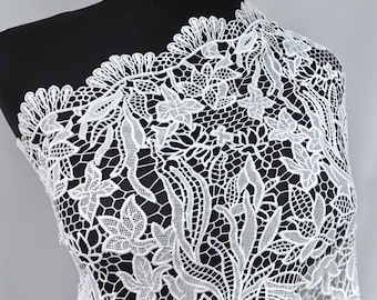 May include: White floral lace fabric draped over a black mannequin. The lace features a detailed pattern of flowers and leaves, with a scalloped edge. The delicate design is visible against the dark background.