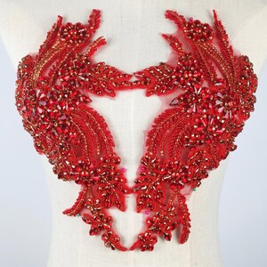 May include: A symmetrical red lace applique featuring intricate floral and leaf patterns. The design is heavily embellished with faceted red crystals, beads, and metallic gold accents, all secured onto a sheer red mesh base for garment decoration.