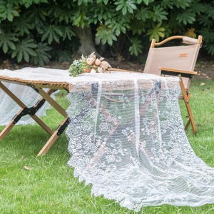 Peut inclure: Une nappe en dentelle blanche à motifs floraux drapée sur une table pliante en bois. Un bouquet de fleurs repose sur la table et une chaise pliante beige est à proximité. La scène se déroule à l'extérieur sur une pelouse.