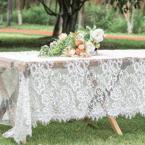 May include: A white lace tablecloth with a floral pattern on a wooden table. A bouquet of white, peach, and green flowers sits on the table. The tablecloth has a scalloped edge and drapes over the table.
