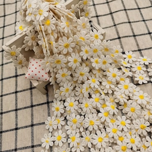 May include: White daisy flower trim with yellow centers. The daisy trim is arranged on a white wooden box with a small red and white polka dot pillow. The background is a black and white checkered pattern.