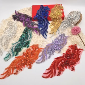 May include: A collection of nine ornate, elongated floral-shaped appliques featuring intricate beadwork and faceted crystals in various colors including red, orange, purple, green, blue, gold, silver, and white, arranged on a white lace fabric.