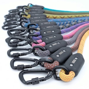 Puede incluir: Una colección de correas para perros de cuerda de colores con clips de metal negro y tapones de goma negra. Las correas están dispuestas en semicírculo con los clips mirando al espectador. Las correas están hechas de diferentes colores, incluyendo amarillo, naranja, rojo, rosa, morado, azul, verde y marrón.