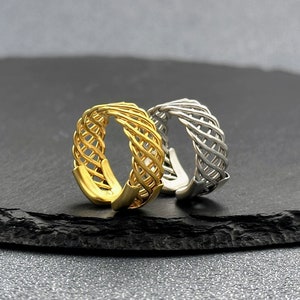 Puede incluir: Dos anillos, uno dorado y otro plateado, con un diseño tejido. Los anillos están sobre una superficie negra.