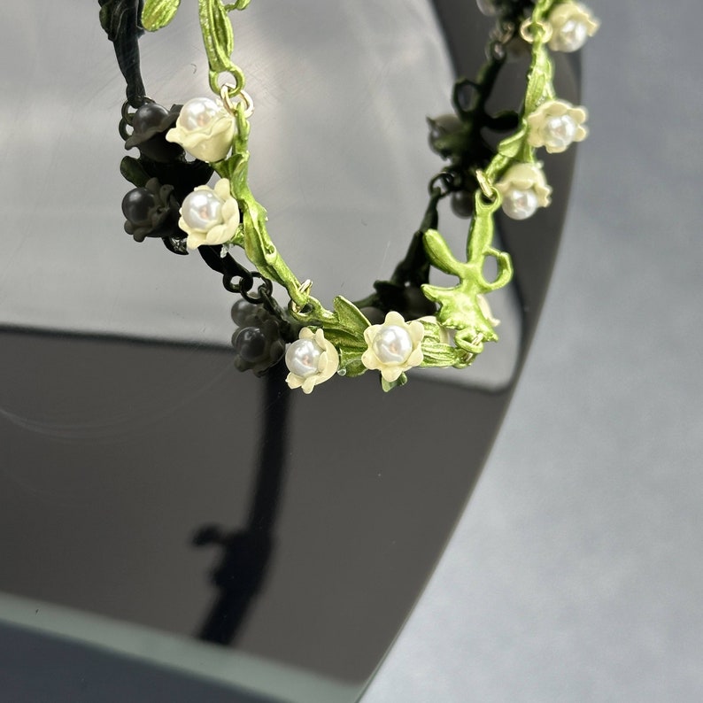 May include: A delicate green and white floral bracelet with pearl accents. The bracelet is made of metal and features a vine design with small flowers and pearls.