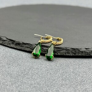 May include: A pair of gold hoop earrings with teardrop-shaped green gemstones. The earrings have a delicate design and are perfect for adding a touch of sparkle to any outfit.