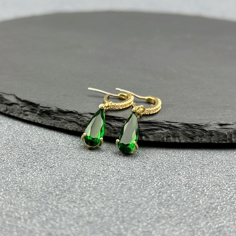 May include: A pair of gold hoop earrings with teardrop-shaped green gemstones. The earrings have a delicate design and are perfect for adding a touch of sparkle to any outfit.