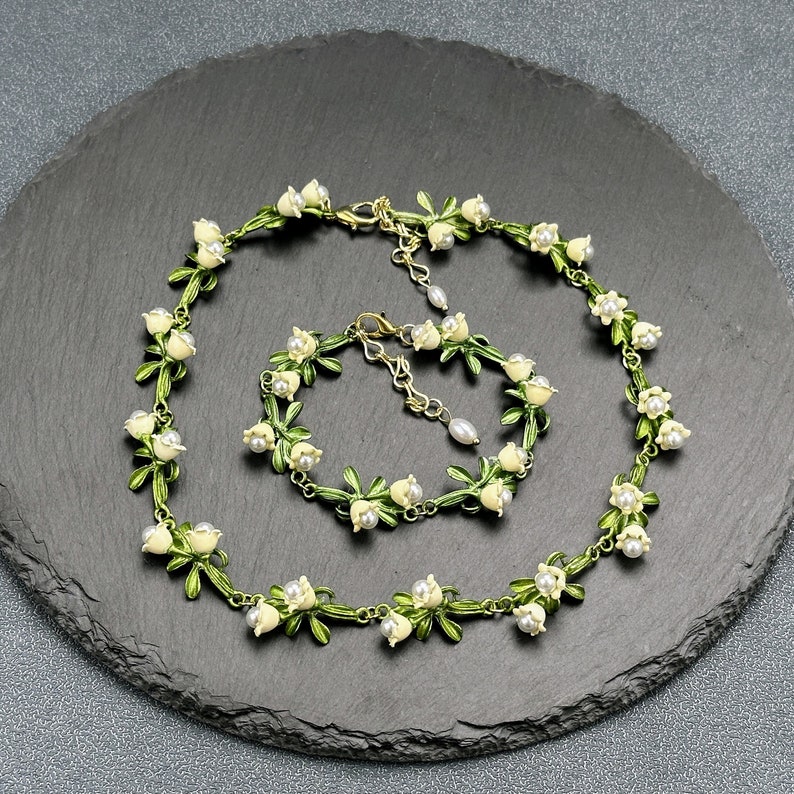 May include: A delicate white flower necklace and bracelet set with green leaves and pearl accents. The jewelry is displayed on a black slate surface.