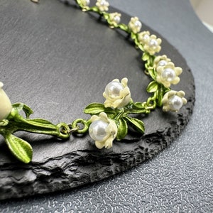 May include: A delicate necklace with a floral design featuring white enamel flowers with pearl accents and green leaves. The necklace is made of metal and has a gold finish.