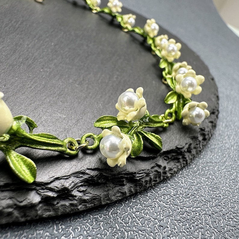 May include: A delicate necklace with a floral design featuring white enamel flowers with pearl accents and green leaves. The necklace is made of metal and has a gold finish.