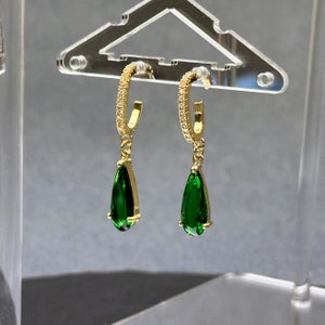May include: A pair of gold hoop earrings with a teardrop-shaped green gemstone dangling from each hoop.