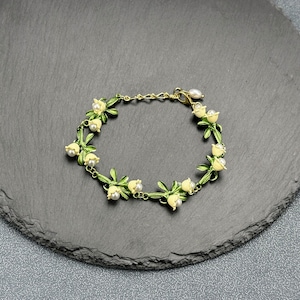 May include: A delicate gold bracelet with a chain and a series of white flower charms with green leaves. The bracelet is on a dark gray background.