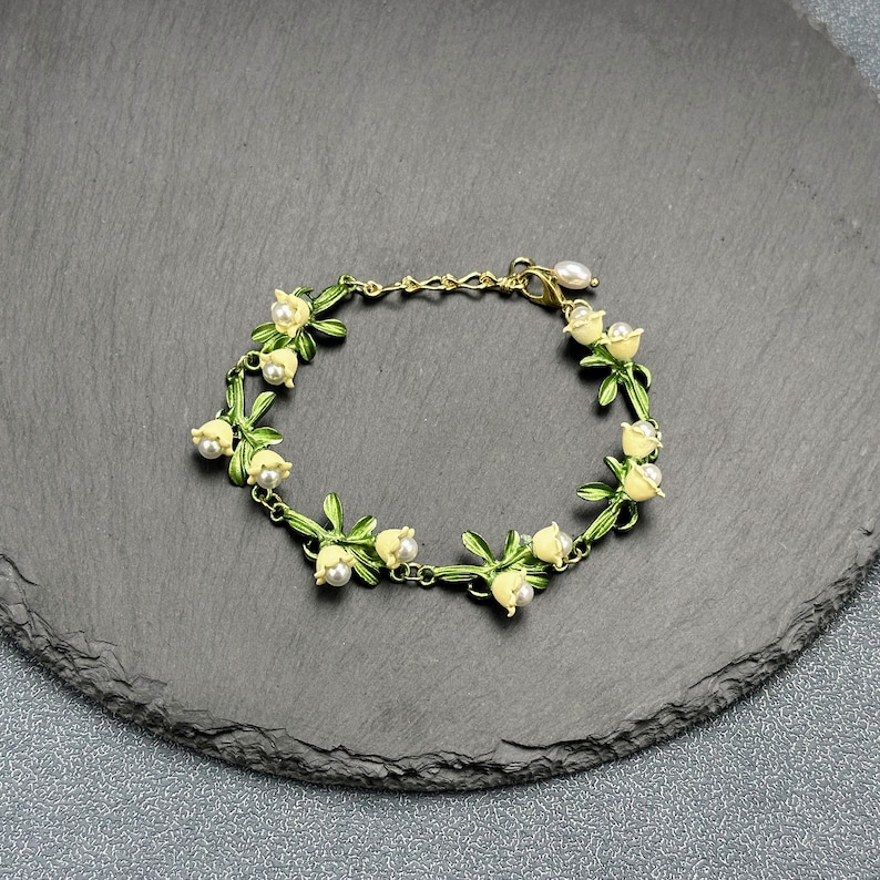 May include: A delicate gold bracelet with a chain and a series of white flower charms with green leaves. The bracelet is on a dark gray background.