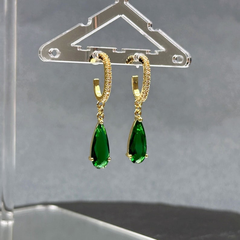 May include: Gold hoop earrings with a teardrop-shaped green gemstone dangling from each hoop. The hoops are encrusted with small clear stones.
