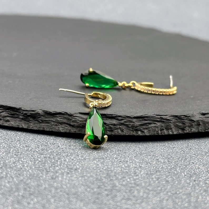 May include: A pair of gold earrings with teardrop-shaped green gemstones and a small row of clear stones around the hoop.