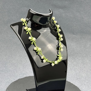 May include: A delicate white and green floral necklace on a black display stand. The necklace features small white flowers with green leaves.