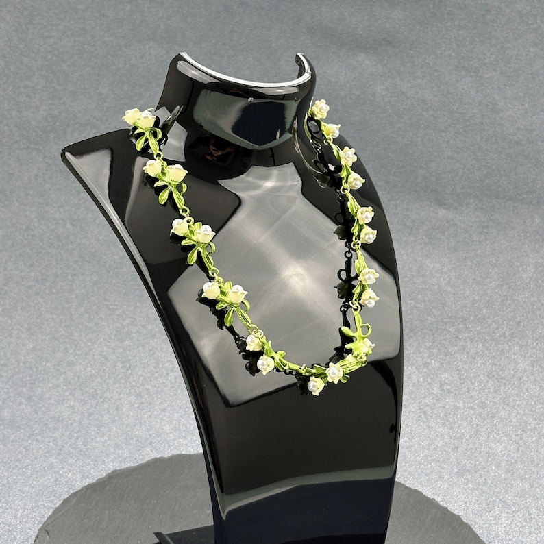 May include: A delicate white and green floral necklace on a black display stand. The necklace features small white flowers with green leaves.