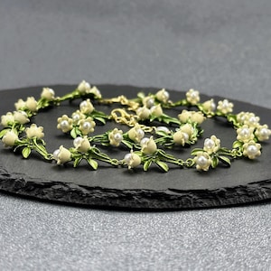May include: A delicate gold chain necklace with white enamel flowers and pearl accents. The necklace features a floral design with green leaves and white blossoms.