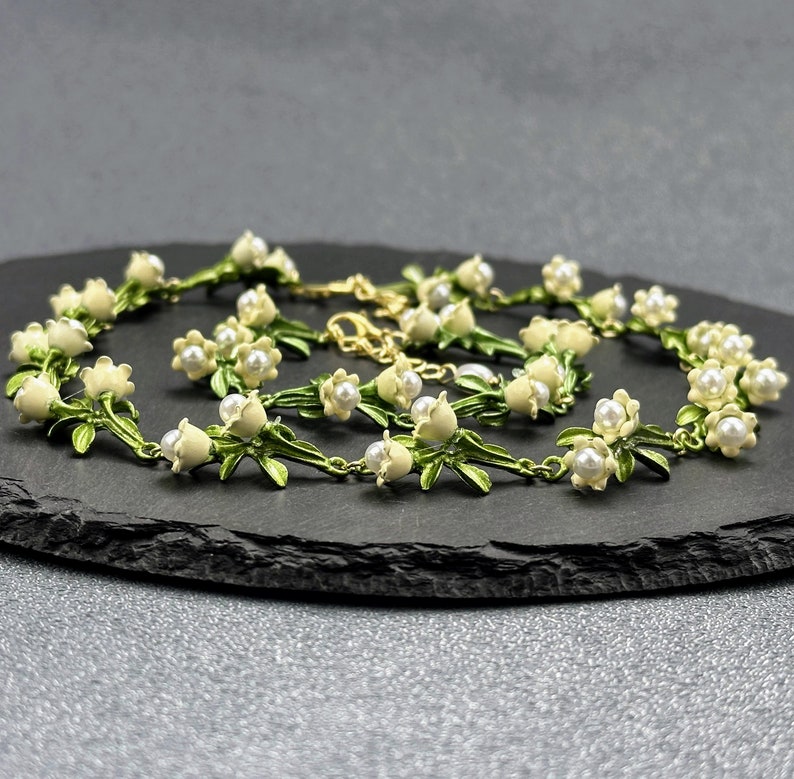 May include: A delicate gold chain necklace with white enamel flowers and pearl accents. The necklace features a floral design with green leaves and white blossoms.